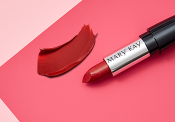 Fotografía de un lápiz labial rojo Mary Kay® sin tapa sobre un fondo rosa junto a una muestra del color del labial.